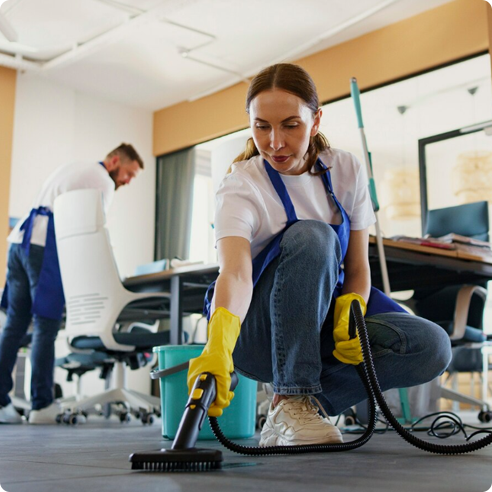 There is a woman kneeling on the ground using a steam cleaner to clean tile while there is a man in the background cleaning a desk.